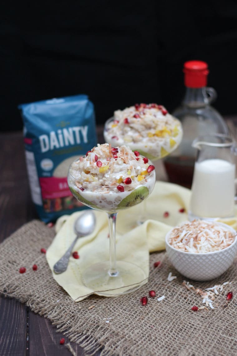 A glass with vegan coconut rice pudding on a table with dainty rice in the background.