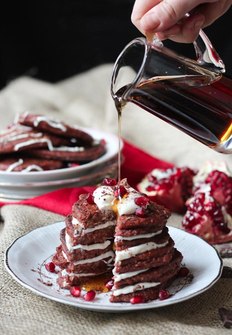 A stack of red velvet pancakes being drizzled with maple syrup.