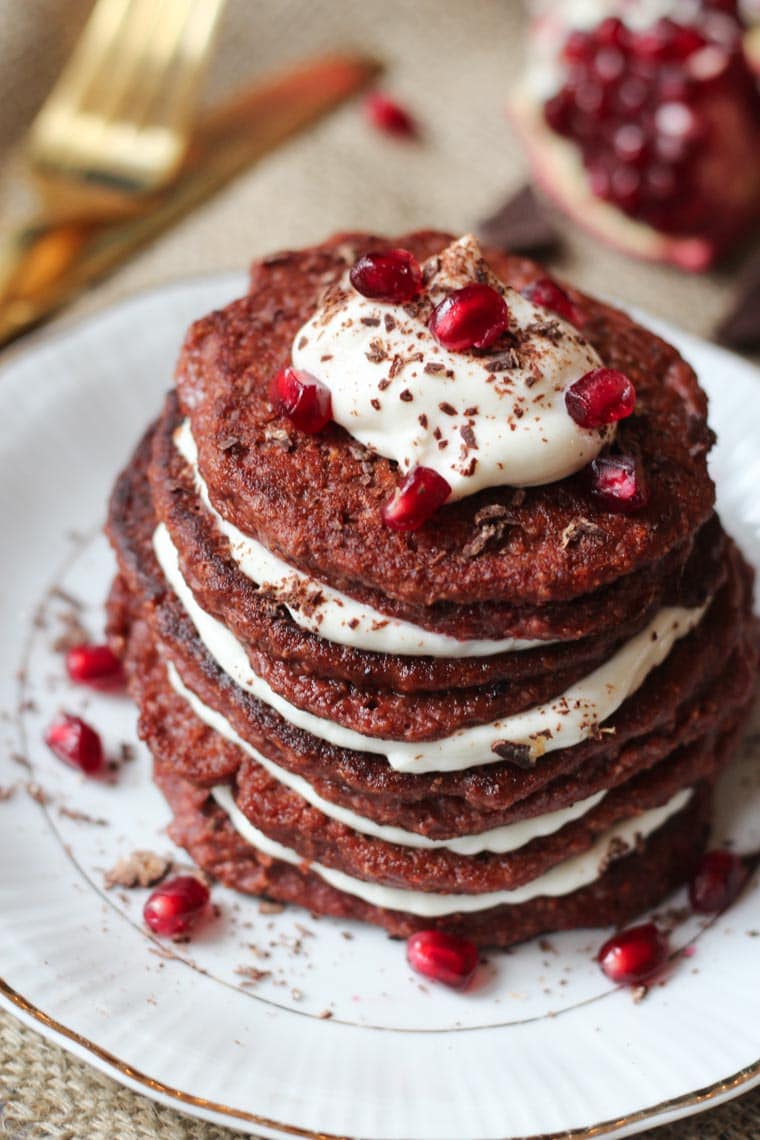 Stack of red velvet pancakes topped with pomegranate seeds, shaved chocolate and cream cheese.
