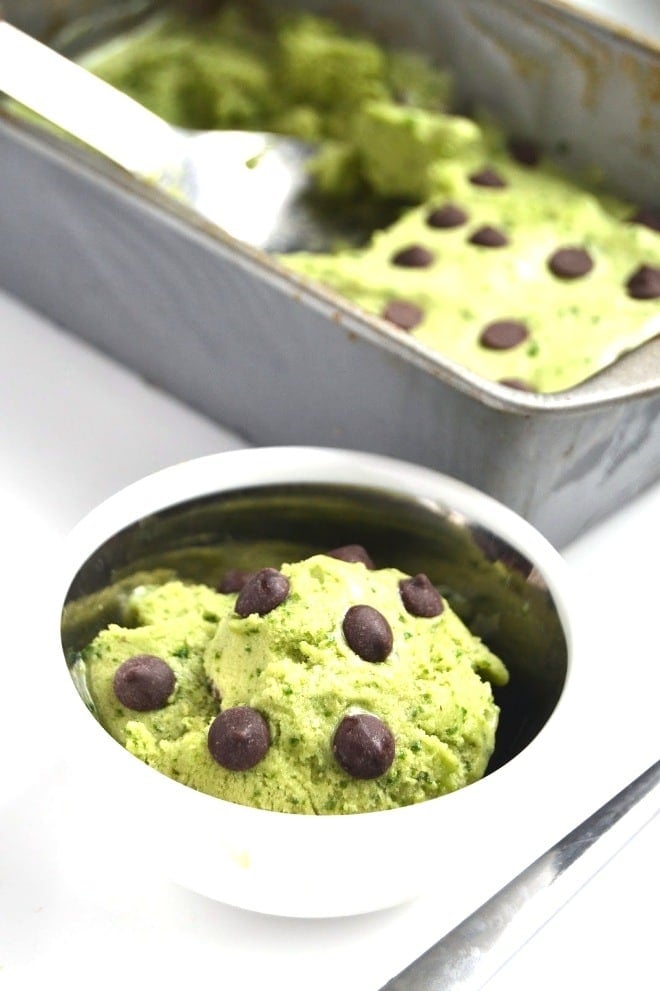 close up of vegan mint chocolate chip protein ice cream garnished with additional chocolate chips served in a white bowl with additional ice cream in a grey tray in the background