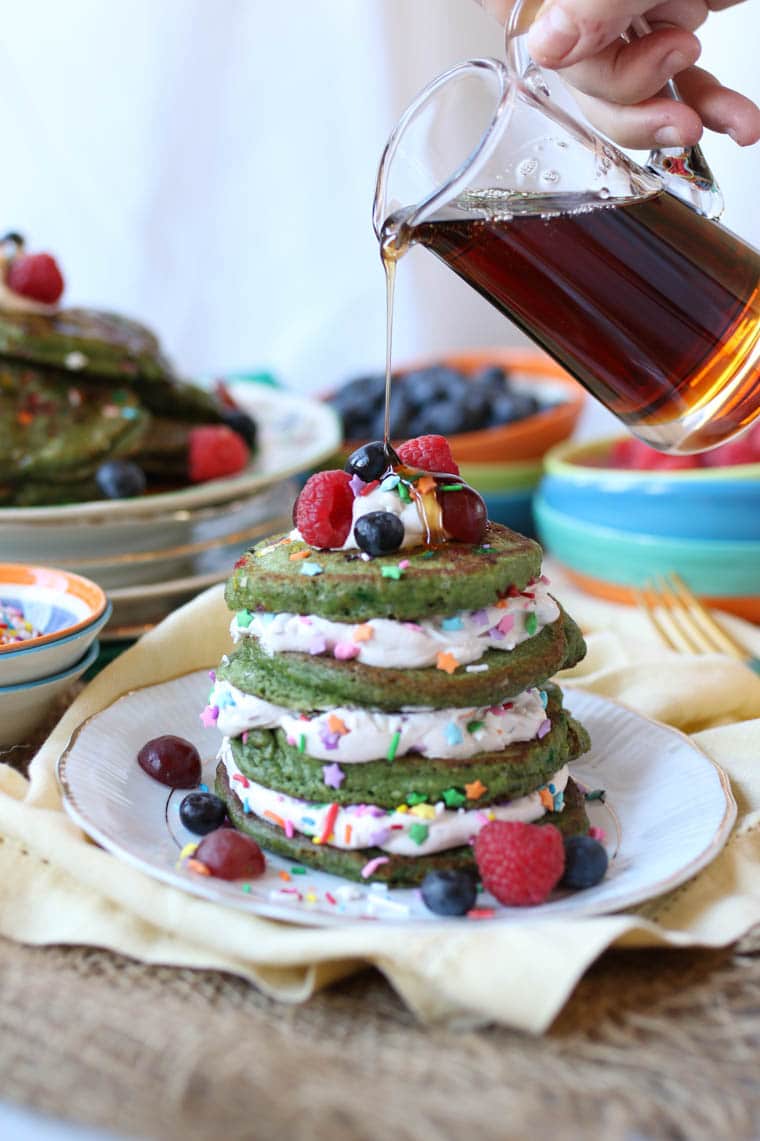 maple syrup being drizzled onto vegan and gluten free protein pancakes on a white plate garnished with whipped cream and berries
