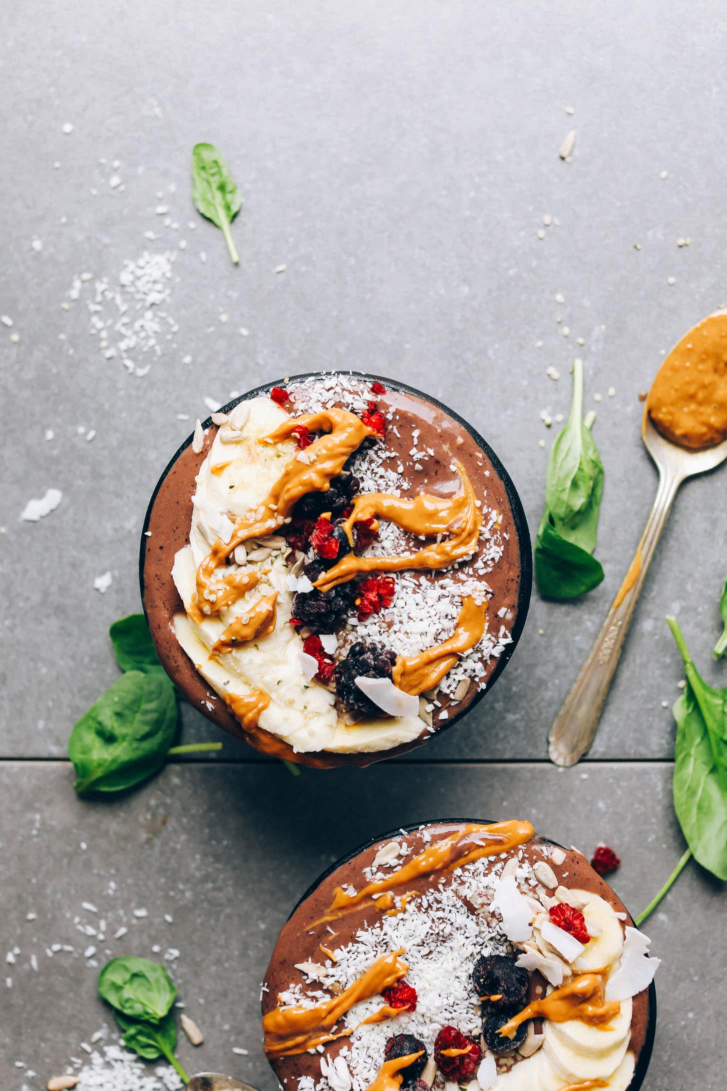 Two smoothie bowls and scattered spinach on a grey surface.