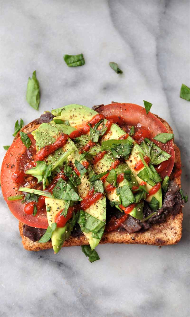 Piece of toast with beans, tomato and avocado on a marble table.