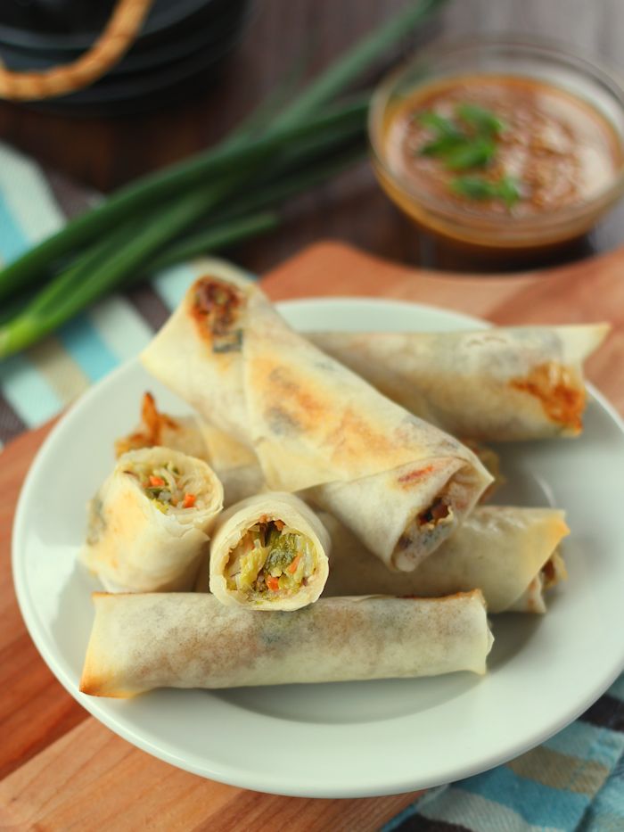 A pile of vegetable spring rolls on a white serving plate with green onions and a dipping sauce bowl in the background.
