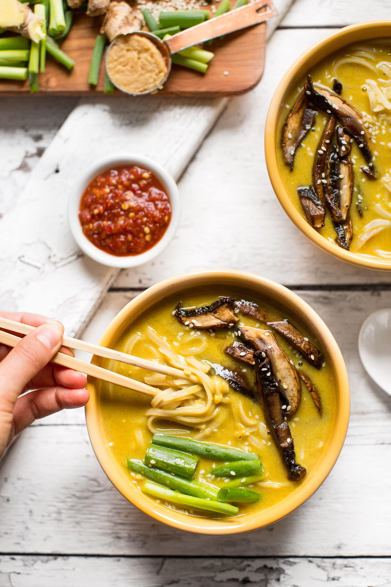 a cream bowl of coconut curry ramen with mushrooms and green onions on top with a pair of chopsticks lifting out some noodles.