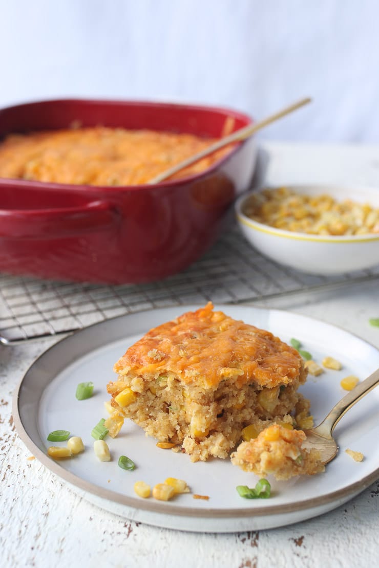 slice of spoonbread on white plate with red casserole dish and corn in the background