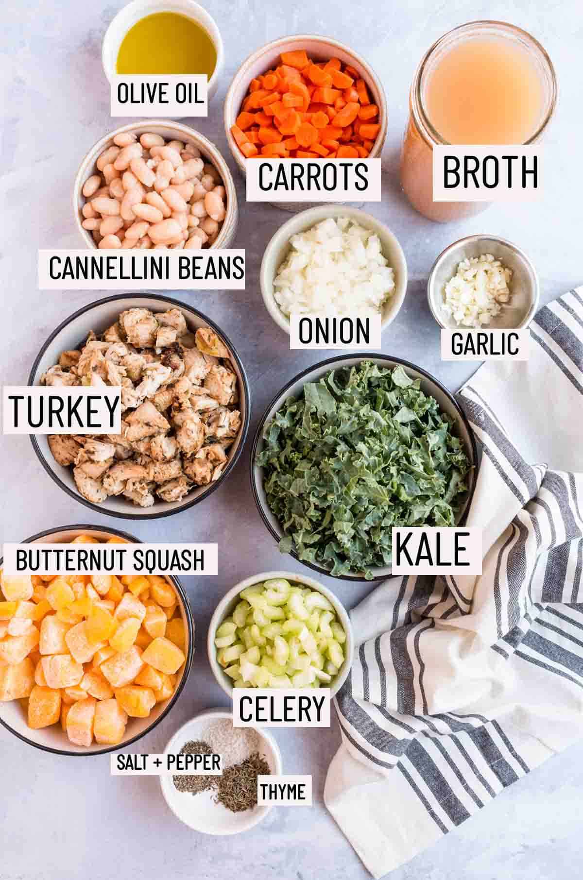 Birds eye view of portioned ingredients to make leftover turkey soup including celery, butternut squash, turkey, cannellini beans, broth, carrots, kale, onion, garlic, and chicken broth. 