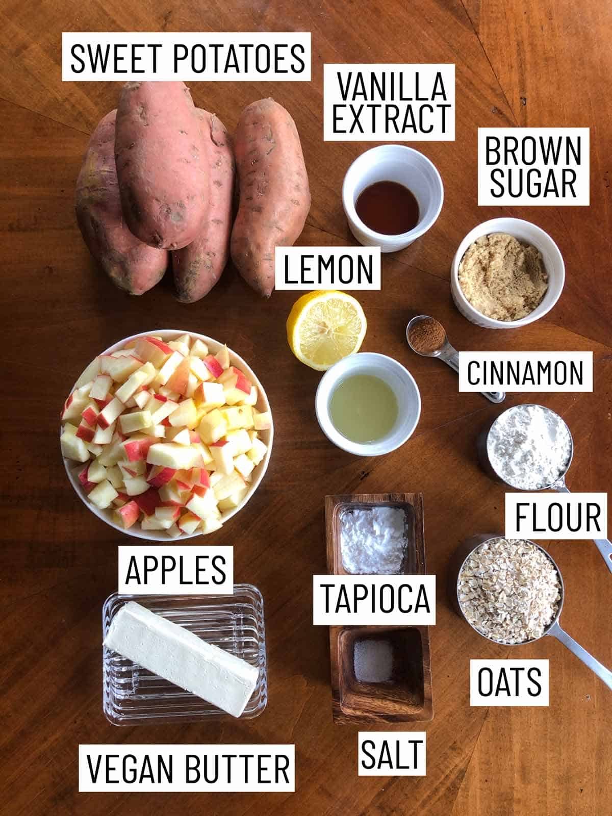 Overhead photo of ingredients containing yams, vanilla extract, brown sugar, cinnamon, lemon, flour, apples, tapioca, oats, salt, and vegan butter.