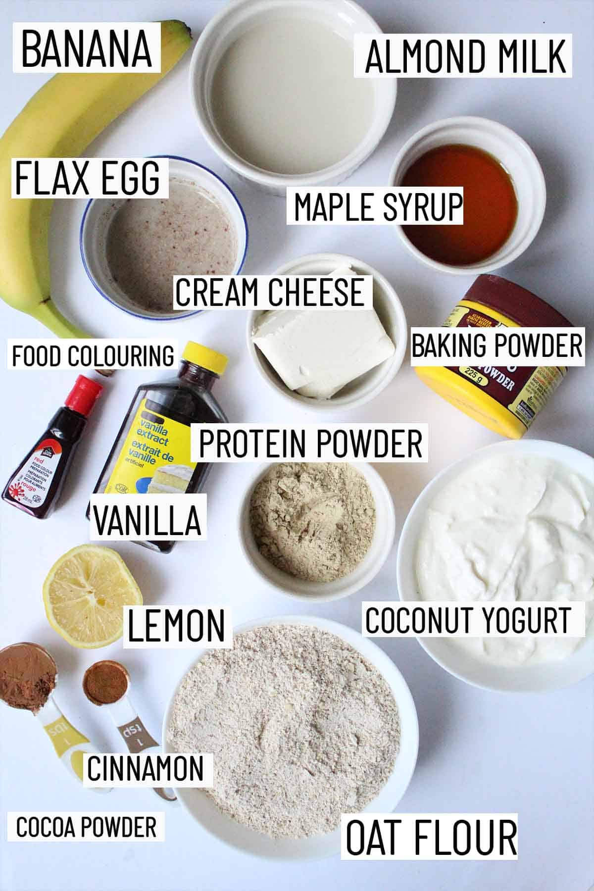 Flat lay image of recipe ingredients including oat flour, cocoa powder, cinnamon, lemon, protein powder, vanilla, food colouring, cream cheese, baking powder, maple syrup, banana, flax egg and almond milk.