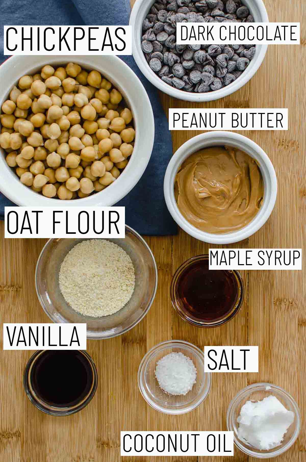 Flat lay image of recipe ingredients including maple syrup, peanut butter, oat flour, vanilla, coconut oil, salt, and chickpeas. 