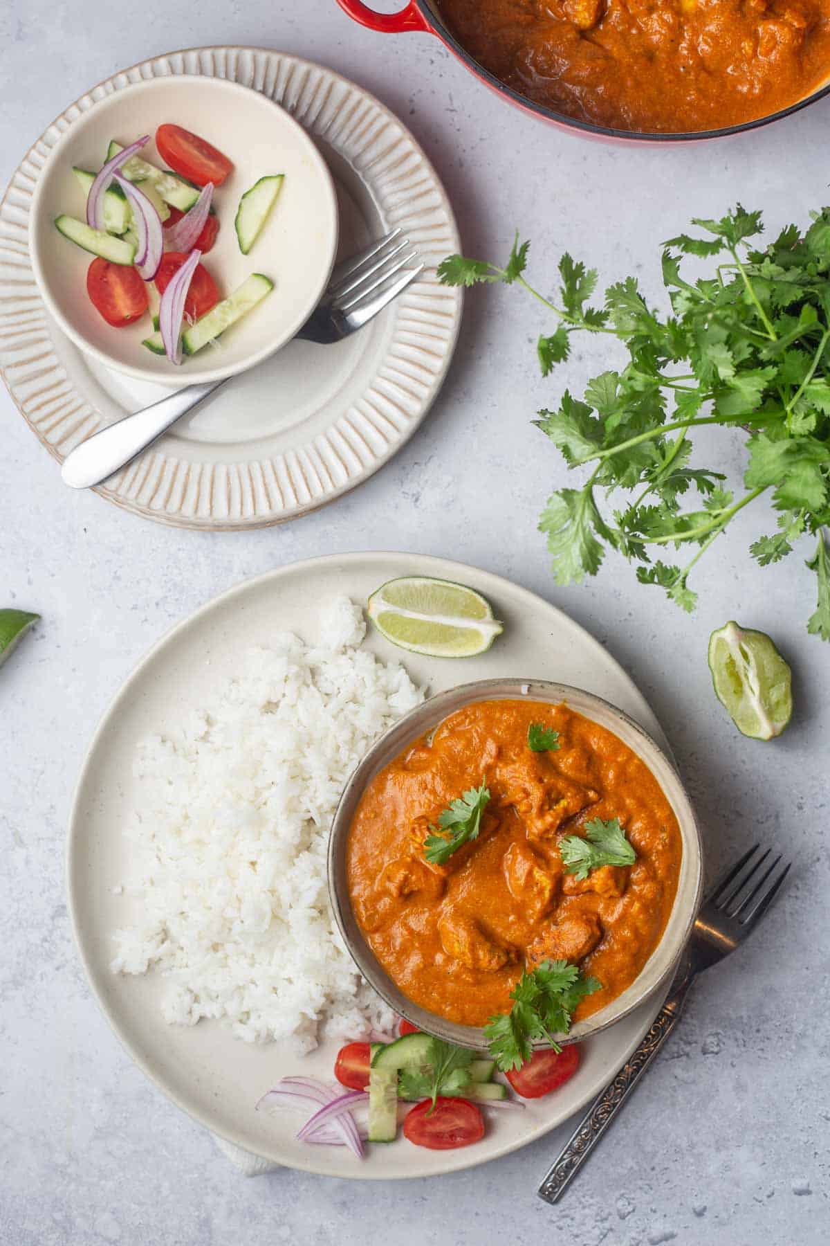 Overhead view of a bowl of butter chicken with a plate of rice beside it with a small bowl of garnish nearby.