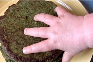 Pinterest graphic of a toddler's hand reaching for the plate of pancakes with text overlay "Baby Led Weaning Green Pancakes Made with Avocado and Spinach."