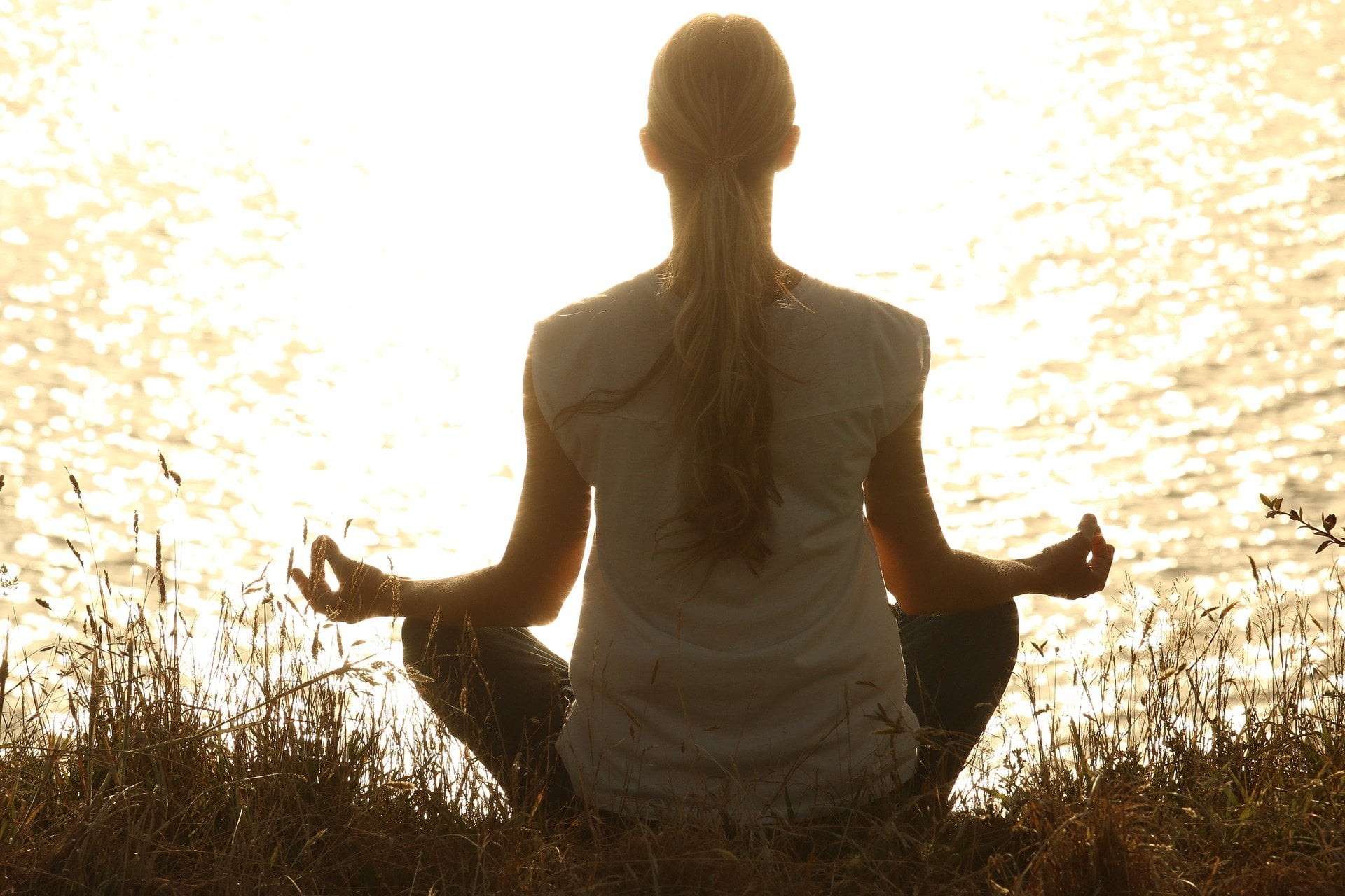 A woman meditating in the grass while facing the water as the sun is setting. 