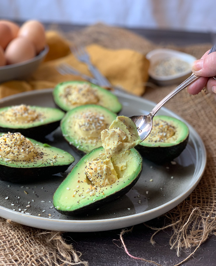 A full shot of a plate of 6 avocado deviled egg halves. A spoon is scooping up some of the filling of the avocado filling in the half closest to the camera.