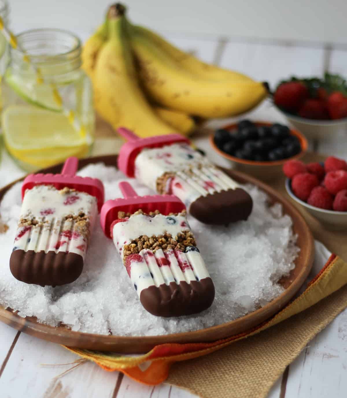 Three banana popsicles on ice with fruits in the background.
